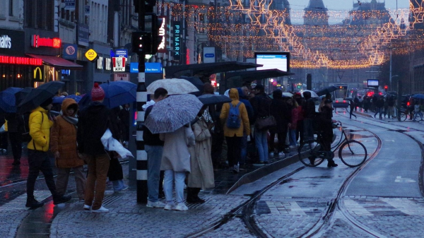 Photograph of people standing at a tram stop with umbrellas up. In the distance the Amsterdam Central Station. It is busy at the tram stop and clearly very rainy weather.