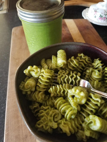 a jar of green pesto and a bowl of radiatore pasta with pesto sauce sitting on a cutting board