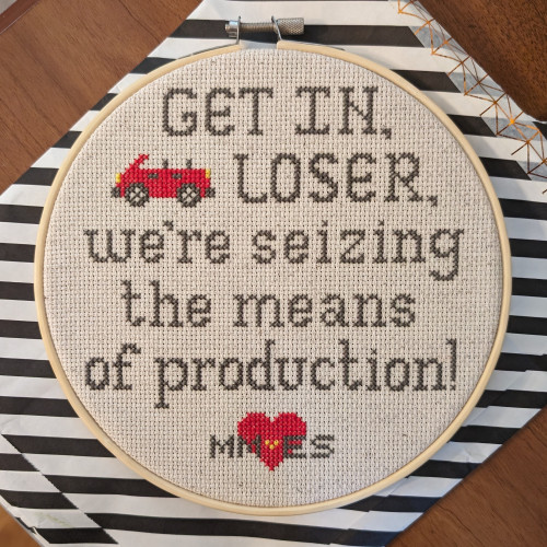 A round embroidery hoop contains a cross stitch design on cream cloth. The words "GET IN, LOSER, we're seizing the means of production!" are stitched in gray thread. A small red convertible and a heart accent the text.  