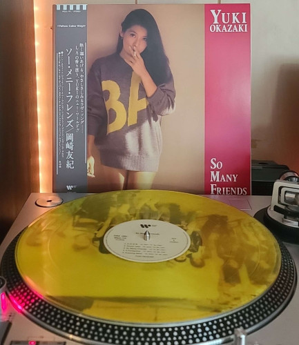 Image shows a turntable with a transparent yellow vinyl record on the platter. Behind the turntable vinyl album outer sleeve is displayed. The front cover shows Yuki Okazaki standing against a wall in an oversized sweater smoking a cigarette. 