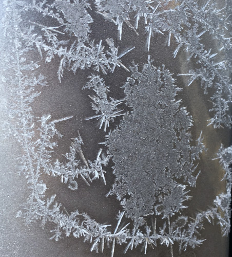 Patches and lines of ice crystals, some quite spiky like cactus, on the gray surface of a car door.