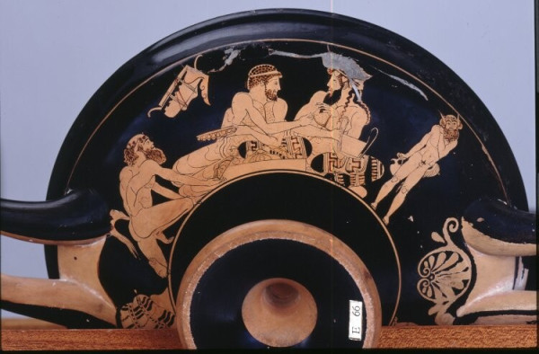Vase painting of a symposion scene with Dionysos and Herakles. They are attended by two satys while they recline on comfortable cushions. Dionysos is holding a kantharos cup and Herakles what looks like a tambourine. Both are bearded and depicted with chest hair.