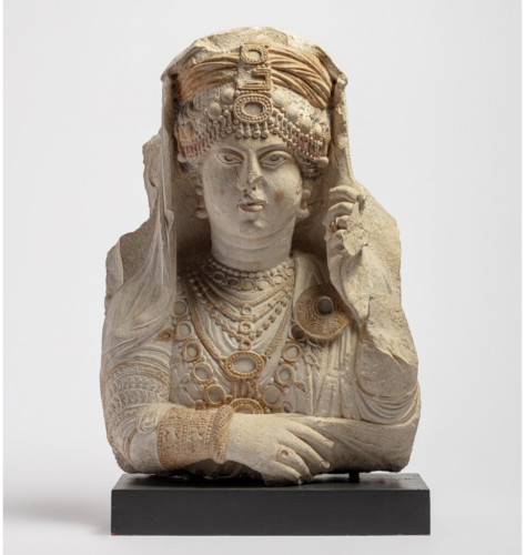 A relief of a woman showing her face, chest, and arms. She crosses her right arm across her body and her left hand holds her veil away from her face. She wears jewellery including necklaces, bracelets, and ornaments over her veil.