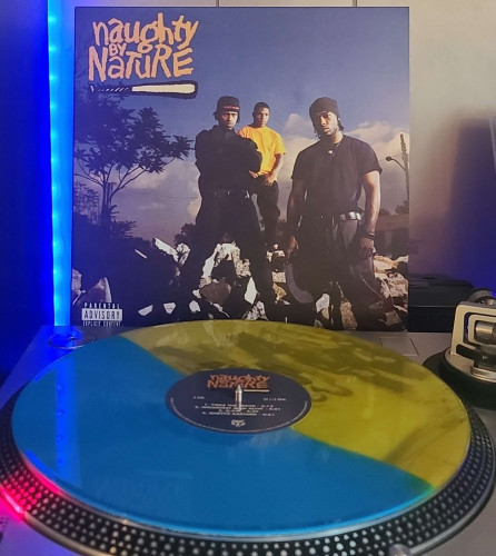 A Blue/Yellow Split vinyl record sits on a turntable. Behind the turntable, a vinyl album outer sleeve is displayed. The front cover shows the 3 members of Naught by Nature standing on rubble with a tree, power lines, clouds, and a blue sky behind them. 