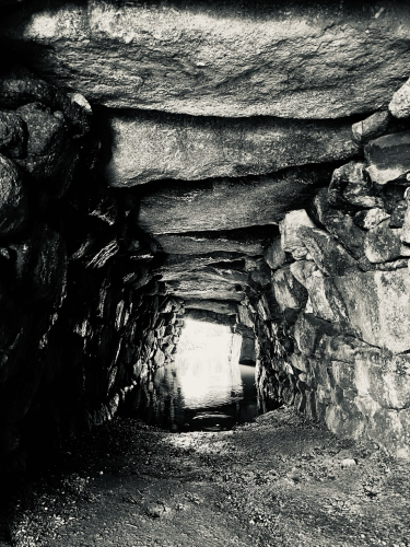 A passageway lined in granite stones along the walls and the ceiling. Rainfall has caused the entrance at the end of the tunnel to have flooded with water.