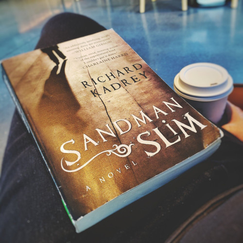 A book titled "Sandman Slim" by Richard Kadrey, perched on a lap beside a to-go cup of coffee.