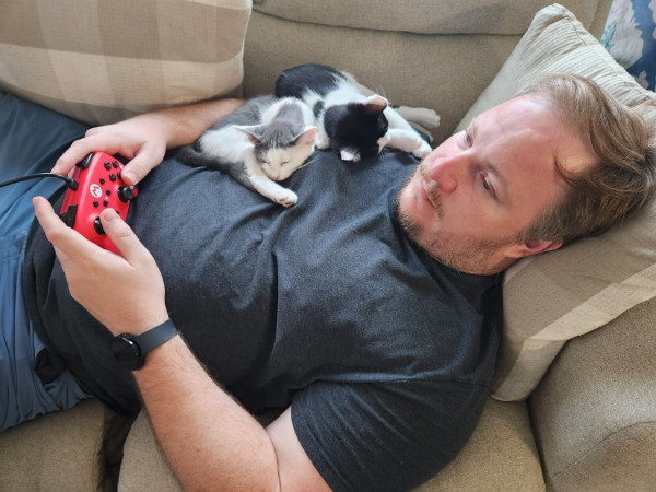 Two kittens laying on a dude laying in a couch. Controller in hand, looking at the tv.