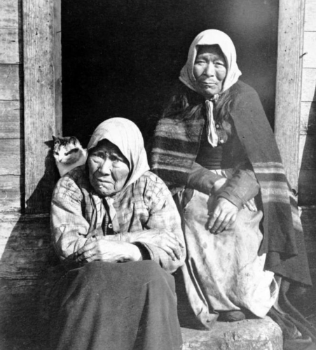 Black and white photo of two older, Alaskan indigenous women sitting in the doorway of a rustic wooden house. They are wearing long skirts, long-sleeved shirts, and head scarves. Behind the should of the woman on the left sits a funny little shorthaired white and tabby cat. Everyone looks quite serious.