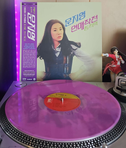 A translucent purple vinyl record sits on a turntable. Behind the turntable, a vinyl album outer sleeve is displayed. The front cover shows Moon Ji-Young looking at the camera with her hands on her hips. 

To the right of the album cover is an anime figure of Yuki Morikawa singing in to a microphone and holding her arm out. 