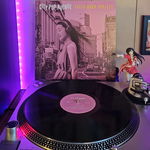 A black vinyl record sits on a turntable. Behind the turntable, a vinyl album outer sleeve is displayed. The front cover shows a Japanese woman in a city wearing headphones and carrying an iced drink in her right hand. 
