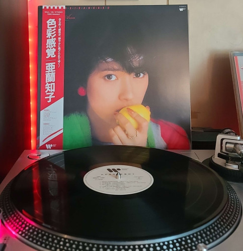 A black vinyl record sits on a turntable. Behind the turntable, a vinyl album outer sleeve is displayed. The front cover shows Tomoko Aran holding a lemon to her lips