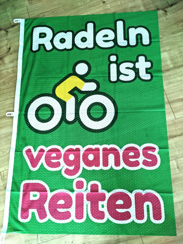 Fahne die auf einem Holzboden liegt. Der Hintergrund ist grün und es steht "Radeln ist veganes Reiten" darauf. In der Mitte ist noch ein stilisierter Radfahrer abgebildet.