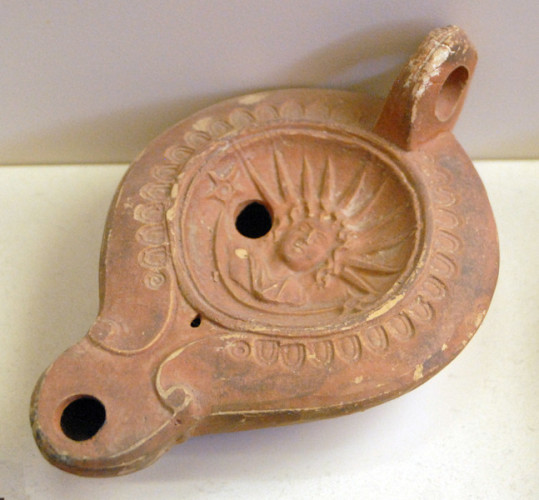 Roman oil lamp decorated with the image of Helios-Sol. The god has chin-length curly hair and two stars are on either side of him. His enormour sun ray crown fills half the circle with his image.