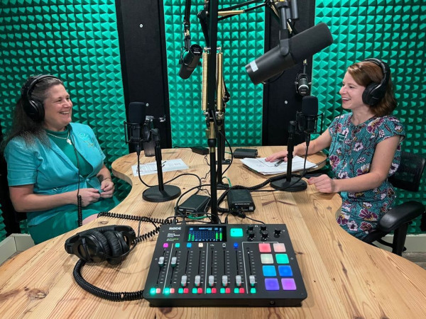 Two women sit smiling at each other in a bright green recording booth. 