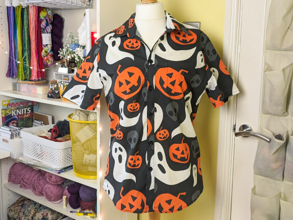 A Halloween shirt on a dressmakers dummy, standing in my craft room next to white shelves full of colourful craft supplies. The shirt is short-sleeved and made out of fabric with a pattern of large, minimalist orange pumpkins and white ghosts on a black background.