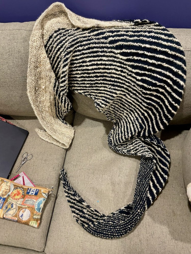 A hand knit shawl is draped over a grey sofa. The shawl is done in stripes of navy blue and off white. Laying on the sofa in the bottom left we see a zippered cloth pouch with knitting tools spilling out on the sofa. 
