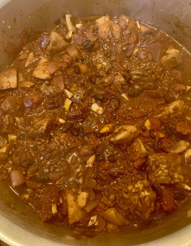 A pot of a stew, red from chiles and tomatoes, that shows mushrooms blocks of tempeh, small piece of lemon, some grain and beans.