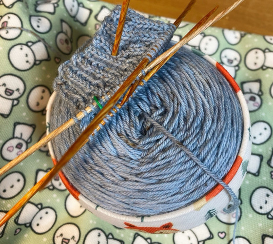 Ball of blue yarn with the cuff of a sock started on the knitting needles. 
