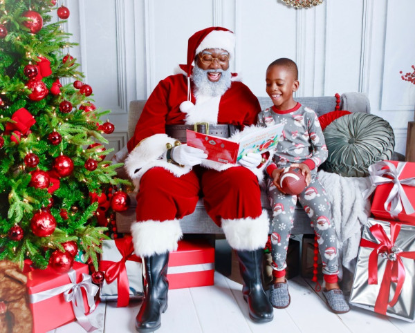 Black Santa and child reading a book together next to a Christmas tree and surrounded by present