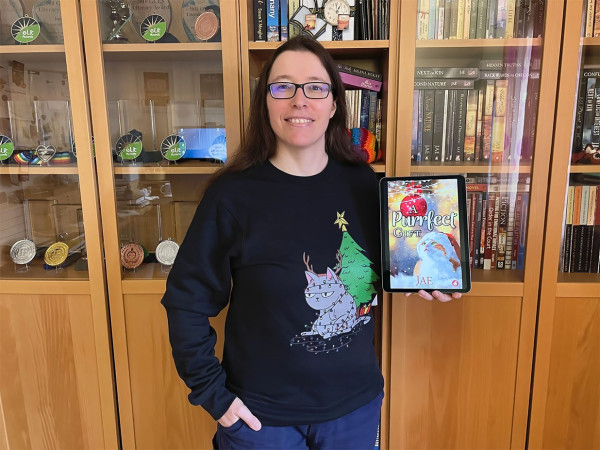 Jae, wearing a Christmas sweater depicting a gray cat, tangled up in a string of Christmas lights. Jae is holding the ebook version of her sapphic Christmas romance novella "A Purrfect Gift", depicting a ginger-and-white cat with a Santa hat. 