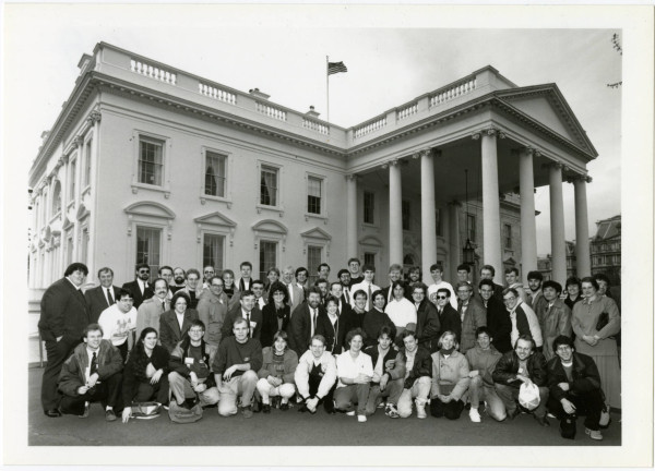 Rectangular black and white image with students and faculty and sponsors in the foreground with the first row on a knee and two rows behind in order of height. The White House is in the background and the group is relatively close to the building on The White House Grounds, and off to the side so an angled perspective of The White House. There are relatively few women in the photo and most appear to be faculty or sponsors or ACM officials or volunteers w/ only several women college students in the photograph.