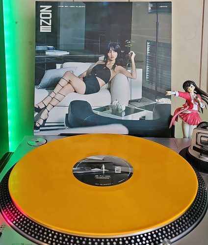 A melon vinyl record sits on a turntable. Behind the turntable, a vinyl album outer sleeve is displayed. The front cover shows Jihyo sitting laid back in a chair

To the right of the album cover is an anime figure of Yuki Morikawa singing in to a microphone and holding her arm out. 
