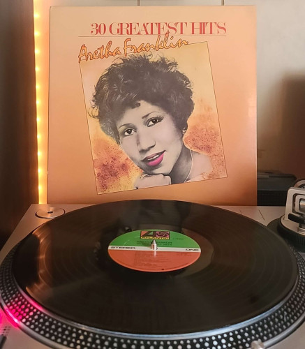 A black vinyl record sits on a turntable. Behind the turntable, a vinyl album outer sleeve is displayed. The front cover shows an artistic portrait of Aretha Franklin
