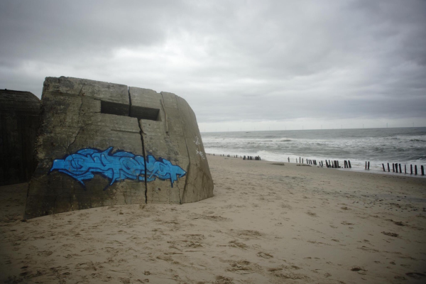 Betonbunker aus dem 2. Weltkrieg am Houvig Strand in Dänemark. Auf der Fassade prangt ein blaues Graffiti in futuristischem Font. Die Szene wirkt wie ein Schauplatz eines Sciencefictionfilmes 