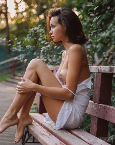 Side view of a pretty lady in a light dress and a gentle romantic pose with her breasts out in open sight sitting on a park bench lustfully gazing towards off camera