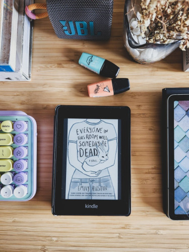 A Kindle displaying the cover of an ebook titled "Everyone in This Room Will Someday Be Dead" by Emily Austin. The Kindle is on a wooden table. Partially seen are a colorful mechanical keyboard and a tablet, plus blue and peach highlighters, a small Bluetooth speaker, a stack of books, and a vase of dried flowers.