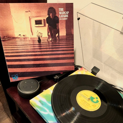Carátula del disco de vinilo de Syd Barret "The Madcap Laughs" al fondo mientras el disco se reproduce en una tornamesa de colores.