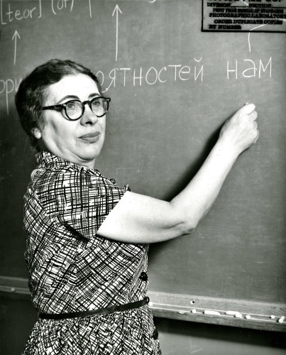 Black and white photo. CBI Image of the Day. Mathematician & pioneering programmer Ida Rhodes in 1959 standing and writing at large chalkboard, instructing, at NBS (there from 1940 to' 71). She is turning away from the board and looking to her right. Portrait orientation photo.