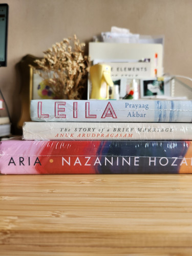 A stack of 3 books on a wooden table, with their spine facing the camera. Behind the stack is an organizing rack with office supplies, a bunch of dried flowers, and a framed artwork.